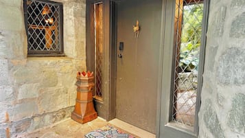 Una entrada de paredes de piedra con una puerta oscura, un pequeño ventanal con reja de metal, un objeto decorativo de madera y un felpudo.
