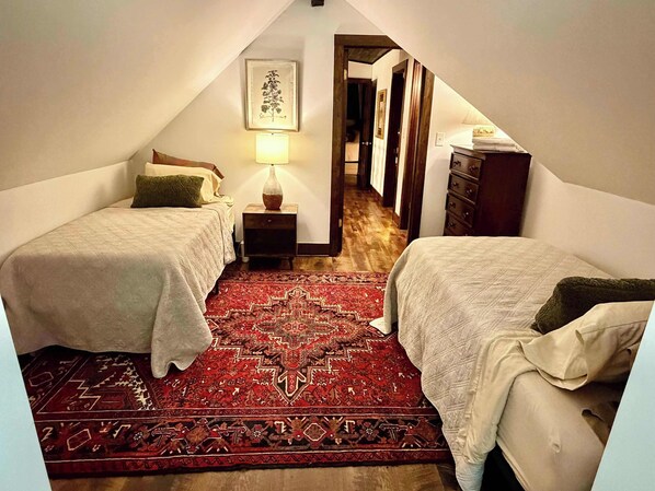 A bedroom with two beds, a red patterned rug, a nightstand with a lamp, and a framed picture on the wall.
