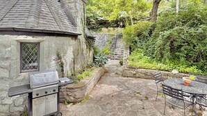 A stone house with a chimney, a barbecue grill, a stone staircase, and a round metal table with chairs.