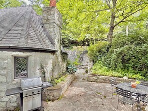 A stone house with a chimney, a barbecue grill, a stone staircase, and a round metal table with chairs.