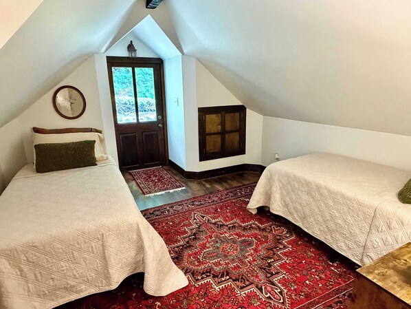A small, triangular attic bedroom with two beds, a large red patterned rug, a wooden door, and a window.