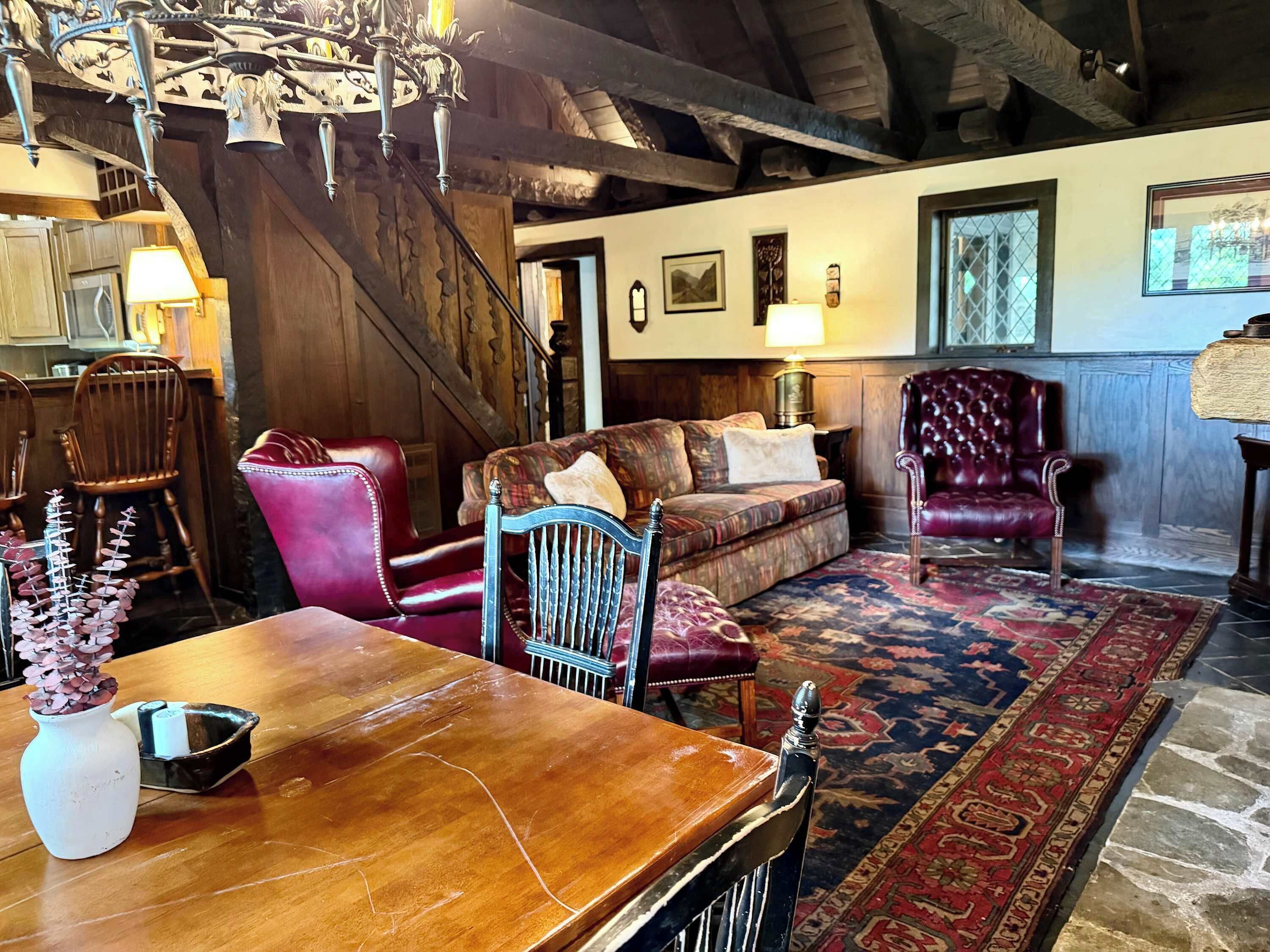 A rustic interior with a wooden dining table, a red leather chair, a patterned sofa, and a chandelier.