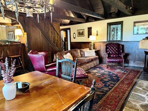 A rustic interior with a wooden dining table, a red leather chair, a patterned sofa, and a chandelier.