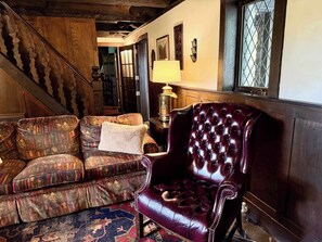 A traditional interior with a wooden staircase, a patterned sofa, a leather armchair, and a lamp.