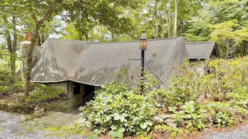 A rustic cabin with a sloped roof, surrounded by dense greenery and a gravel path.