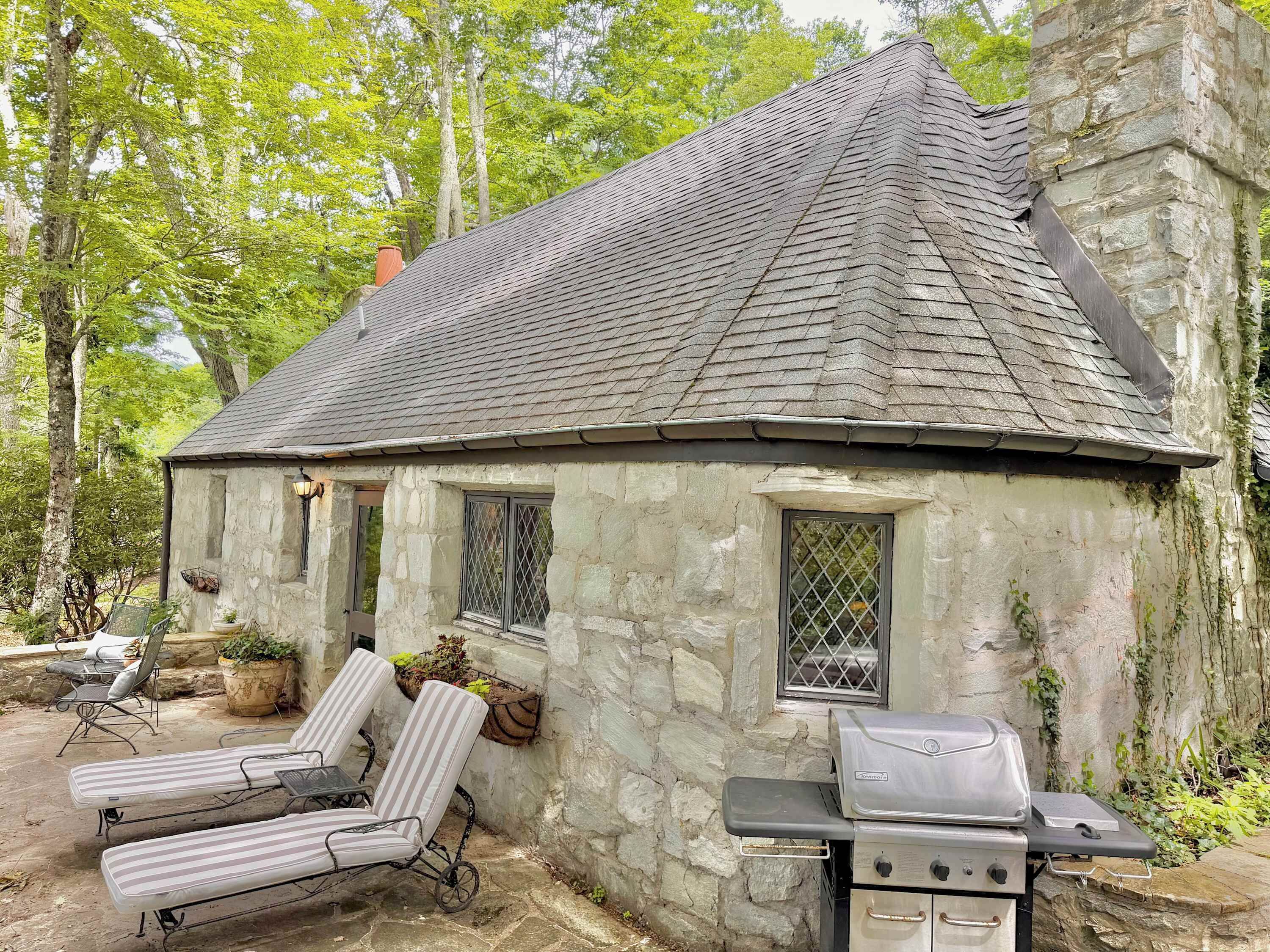 A stone cottage with a chimney, a grill, and two lounge chairs.