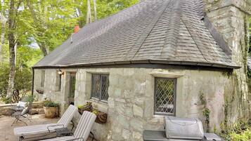 A stone cottage with a chimney, a grill, and two lounge chairs.