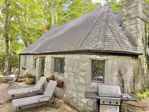 Une petite maison en pierre avec une cheminée, un barbecue et deux fauteuils de jardin.