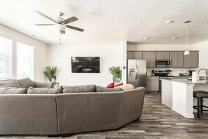 A modern living room with a sectional sofa, a ceiling fan, a kitchen with a microwave, and a dining area.