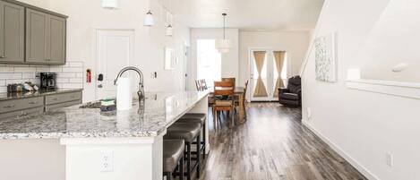 A modern kitchen with a marble countertop, bar stools, and a dining area with a wooden table and chairs.