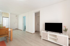 Un salon moderne avec un téléviseur à écran plat posé sur un meuble blanc, une table à manger en bois avec des chaises, et un couloir menant à une autre pièce.