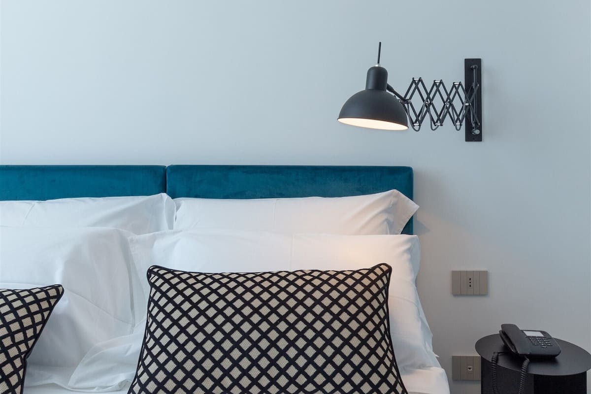 Een bed met witte lakens, een blauw hoofdeinde en gestreepte kussens. Een zwarte bureaulamp is aan de muur naast het bed bevestigd.
