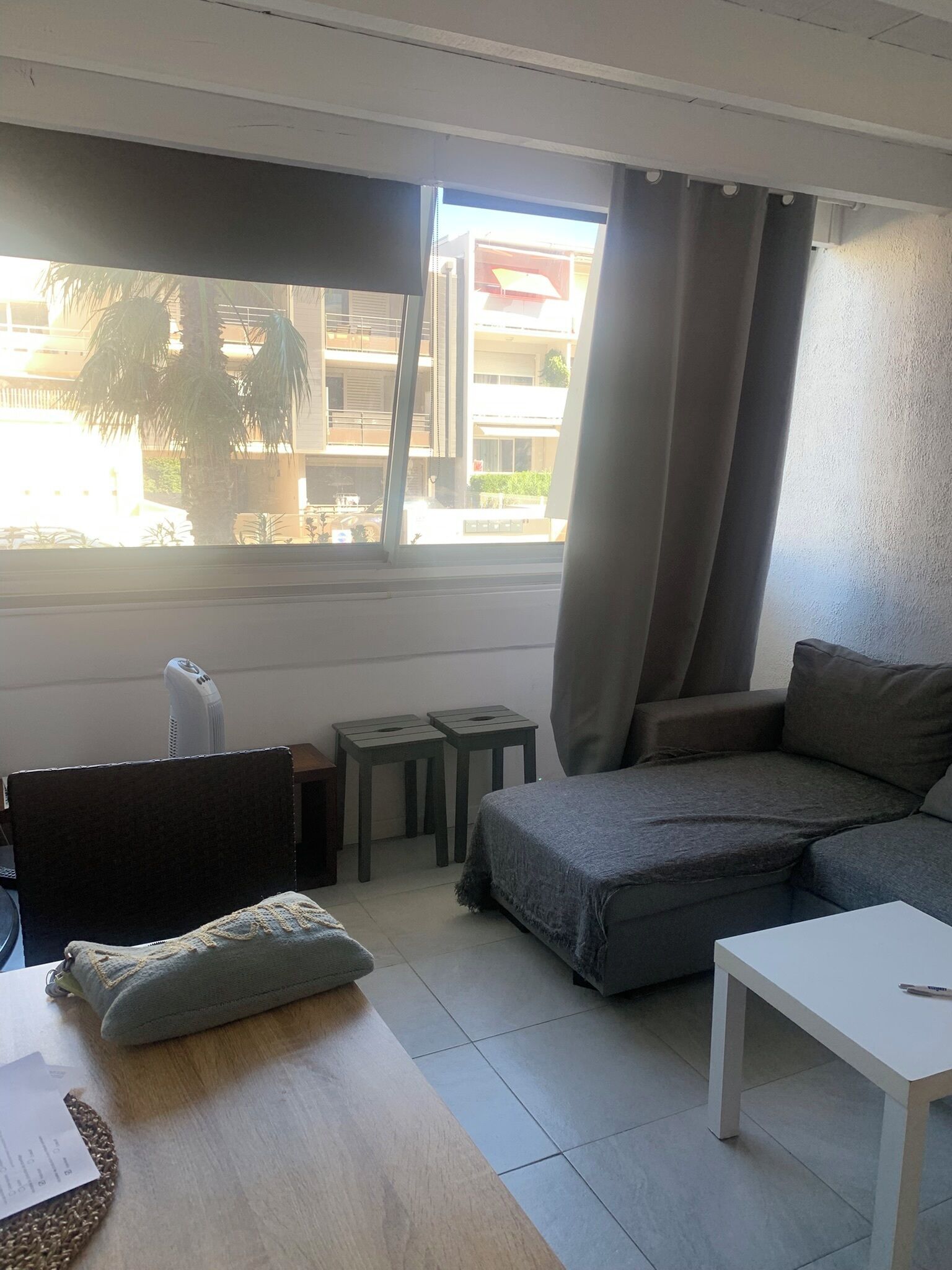 Ein Wohnzimmer mit einer Couch, einem kleinen Tisch, einem Sessel, einem Fenster mit Vorhängen und Blick auf Gebäude.