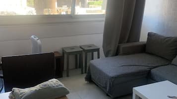Ein Wohnzimmer mit einer Couch, einem kleinen Tisch, einem Sessel, einem Fenster mit Vorhängen und Blick auf Gebäude.