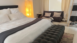 Ein ordentlich eingerichtetes Schlafzimmer mit einem großen Bett, einer dunkel gepolsterten Bank, einem Nachttisch und einer Lampe.