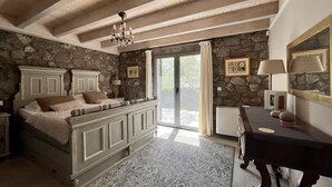A bedroom with a stone wall, a bed with a headboard, a chandelier, a large window, and a nightstand. - Aegean Retreat for 6 Pool & Evia Views Near Athens (Chalcis)