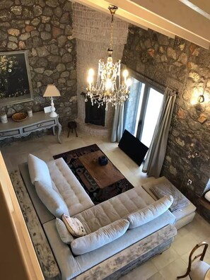 A living room with a stone wall, a chandelier, a sofa, a coffee table, and a flat-screen TV. - Aegean Retreat for 6 Pool & Evia Views Near Athens (Chalcis)
