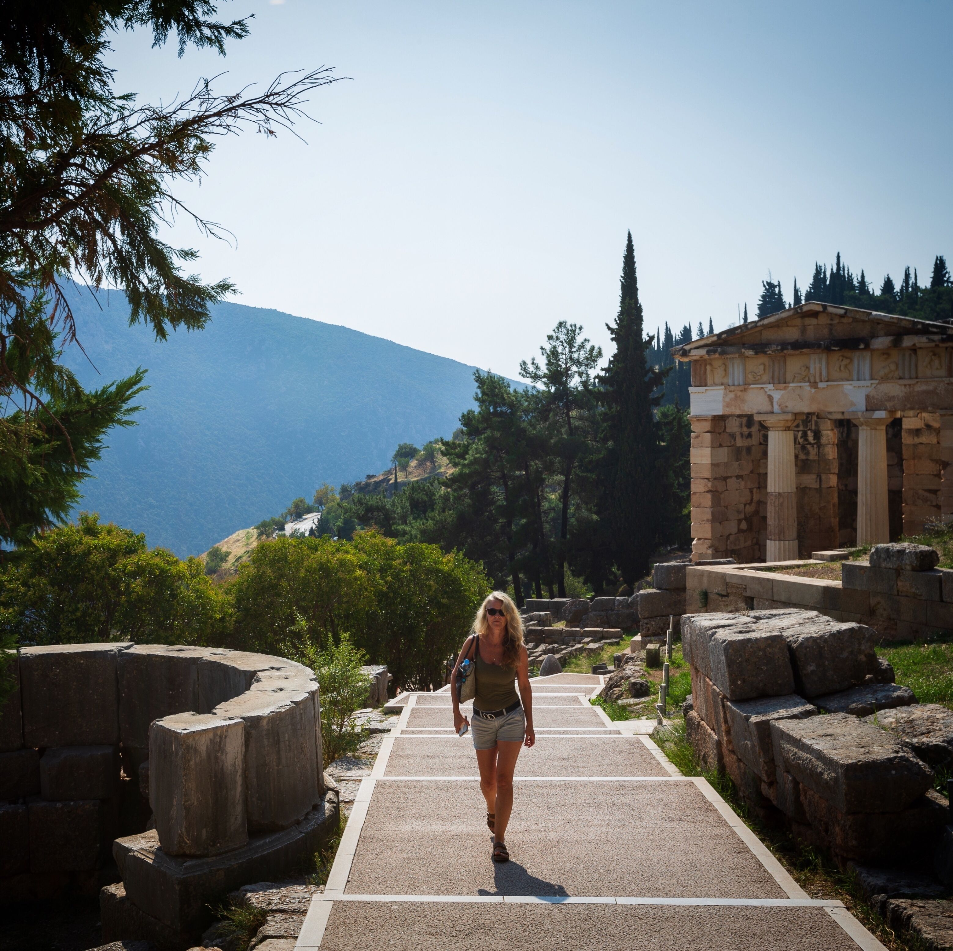 A woman walking on a paved path surrounded by ancient ruins and trees, with a mountain in the background.