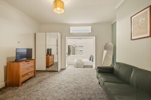 Een kamer met een televisie op een houten dressoir, een groene bank, een grote spiegel en een raam met uitzicht op een slaapkamer.