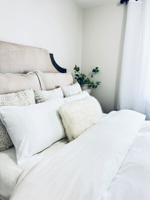 A neatly made bed with white bedding and multiple pillows, a wall-mounted headboard, and a potted plant in the corner. - Cozy 2-bedroom house. Experience Sarasota Living Near America’s Top-Rated Beach (Sarasota)