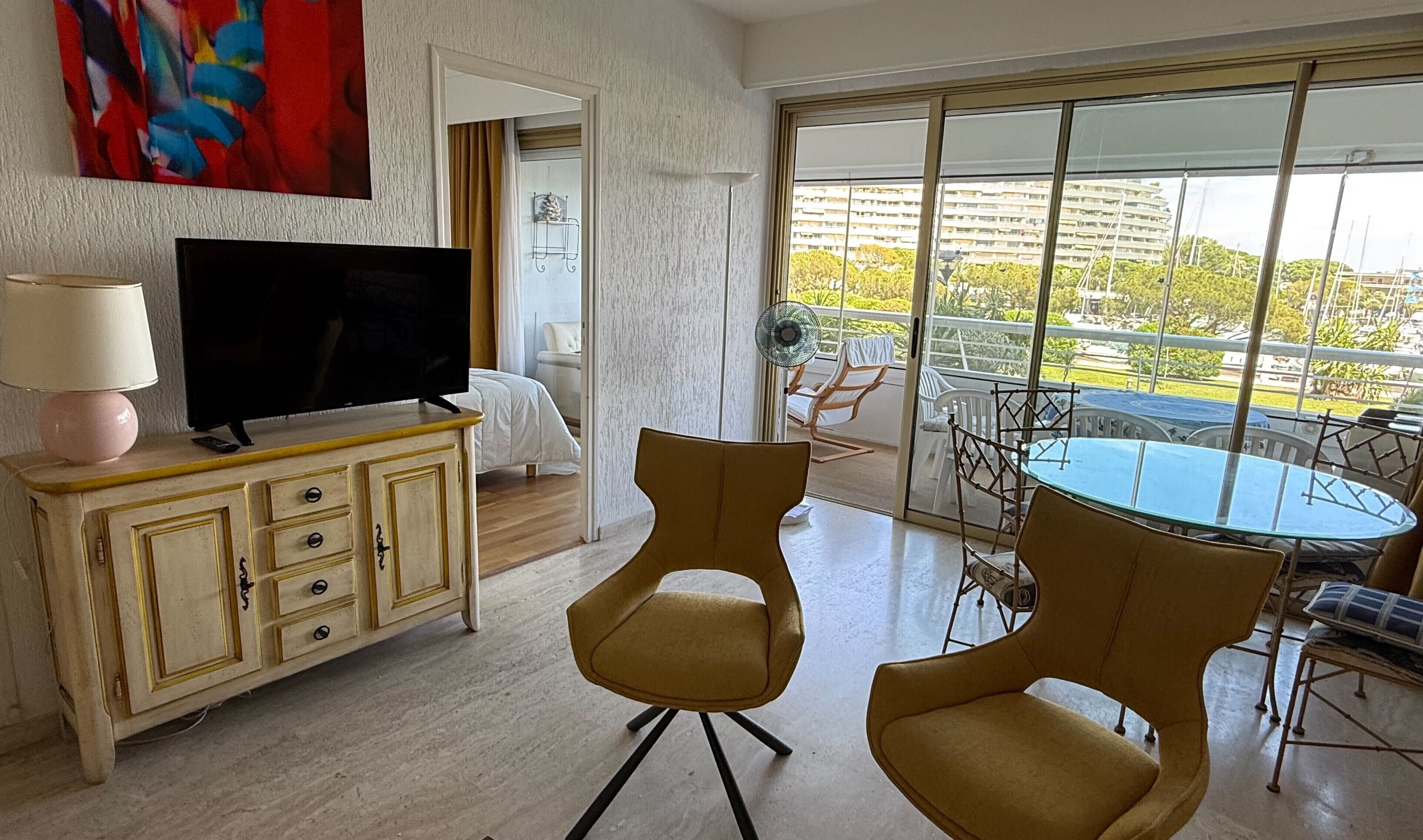 Une chambre d’hôtel équipée d’une télévision, d’un coin repas avec des chaises et une table, d’une grande fenêtre donnant sur des immeubles et d’un tableau accroché au mur.