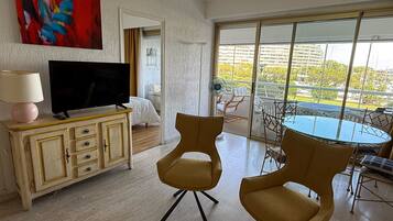Une chambre d’hôtel équipée d’une télévision, d’un coin repas avec des chaises et une table, d’une grande fenêtre donnant sur des immeubles et d’un tableau accroché au mur.