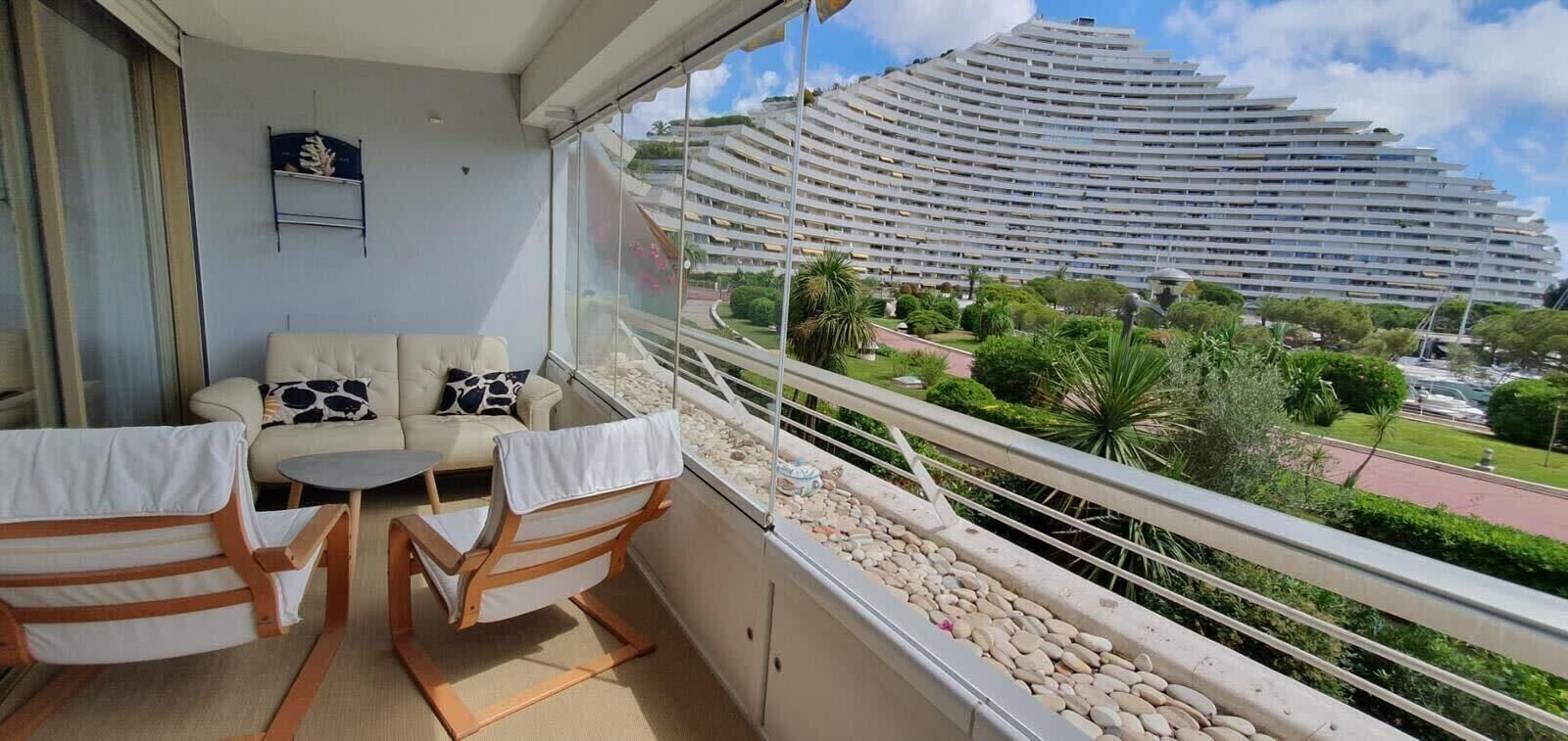 Un balcone con divano e sedie, vista su un edificio moderno e area verde con alberi e una strada.