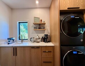 Une cuisine moderne avec des meubles en bois, un four encastré, une machine à café et une fenêtre donnant sur un paysage verdoyant.