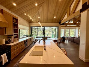 A modern kitchen with a large island, wooden ceiling beams, and large windows offering views of the outdoors.