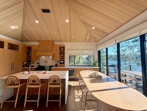 A modern kitchen with wooden cabinets, a central island with bar stools, and large windows offering a view of the outdoors.