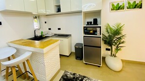 Una cocina moderna con armarios blancos, microondas integrado y un refrigerador pequeño. Hay una isla blanca con taburetes, una planta en maceta y un aplique de pared.
