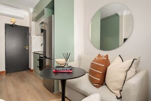 A modern living room with a round mirror, a sofa with patterned cushions, a small table with books, and a kitchen area visible through a glass partition.