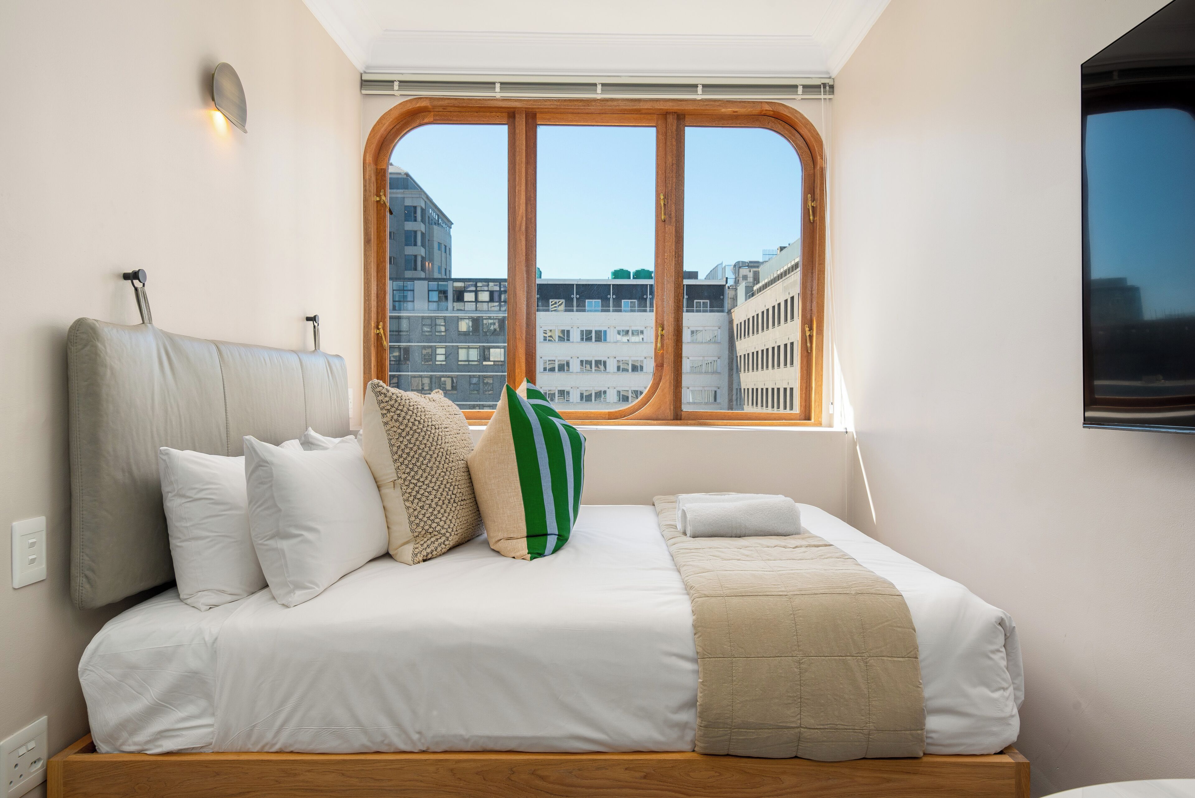 A bed with a headboard, pillows, and a blanket, a window with a view of buildings, and a mounted TV.
