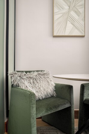 Ein grüner, gepolsterter Sessel mit einem flauschigen weißen Kissen, ein runder Tisch und ein gerahmtes, abstraktes Kunstwerk an der Wand.