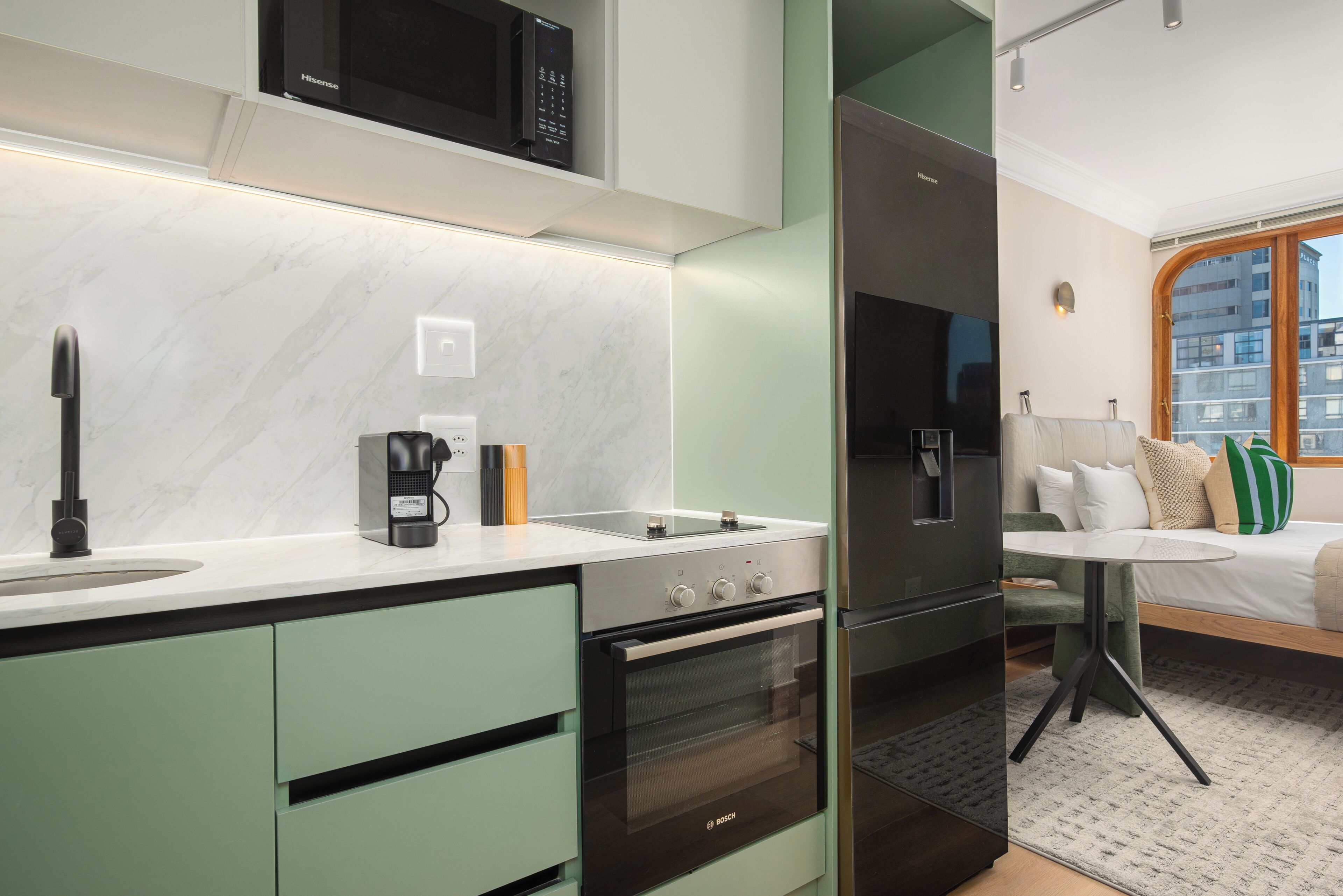 A modern kitchen with a microwave, stove, and oven, a small dining area with a table, and a bed with a view of the city.