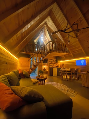 Una habitación estilo loft acogedora con una escalera de madera, un sofá con cojines, un comedor y un televisor.