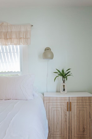 Un dormitorio con cama, una mesita de noche con una planta, una lámpara de pared y un ventanal con persianas.