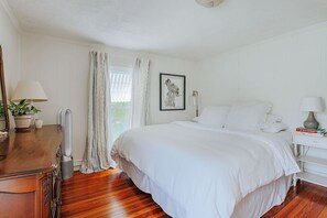 Une chambre avec un plancher en bois, un lit recouvert de draps blancs, une table de chevet avec une lampe, une plante en pot et un tableau encadré accroché au mur.