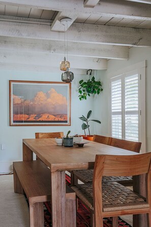 Uma área de refeições com uma mesa de madeira, cadeiras, uma planta pendente e um quadro na parede.