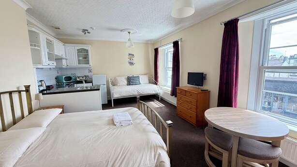 A hotel room with a bed, a small desk with a TV, a dining table, a kitchenette, and a large window. - Belgravia Holiday Apartments No 4 (Torquay)