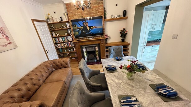 Una sala de estar con un sofá de cuero marrón, un sillón gris, una mesa de comedor de mármol adornada con servilletas y platos azules, un televisor en una pared de ladrillo y una estantería llena de libros y objetos decorativos.