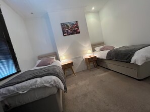 A bedroom with two beds, a nightstand with a lamp, a wall-mounted picture, and a window with blinds. - PITTVILLE HOUSE 
Space and comfort in the beautiful regency town of Cheltenham. (Cheltenham)