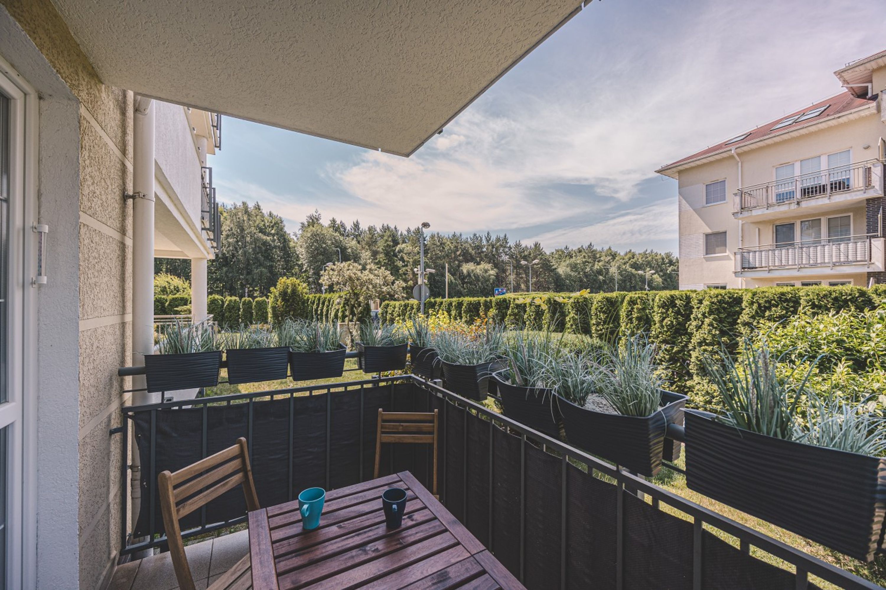 A balcony with a table and chairs, a view of a well-maintained garden, and a building with balconies in the background.