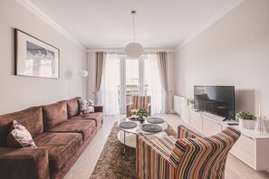 Una sala de estar con un sofá marrón, un sillón a rayas, una mesa de centro redonda y un televisor sobre un mueble blanco.