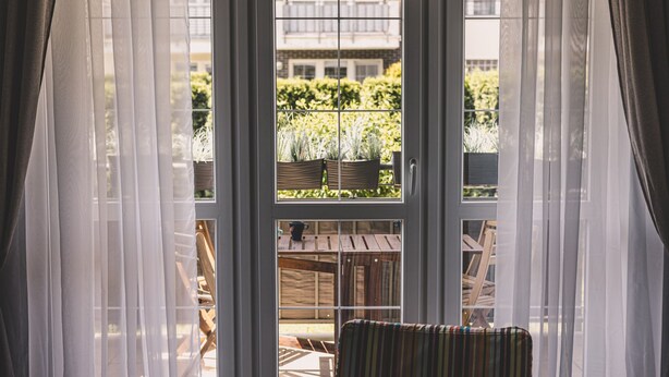 A room with a large window, a chair, and a view of a balcony with plants and outdoor furniture. - Setas Apartment Osiedle Ba tyk Taras (Kołobrzeg)