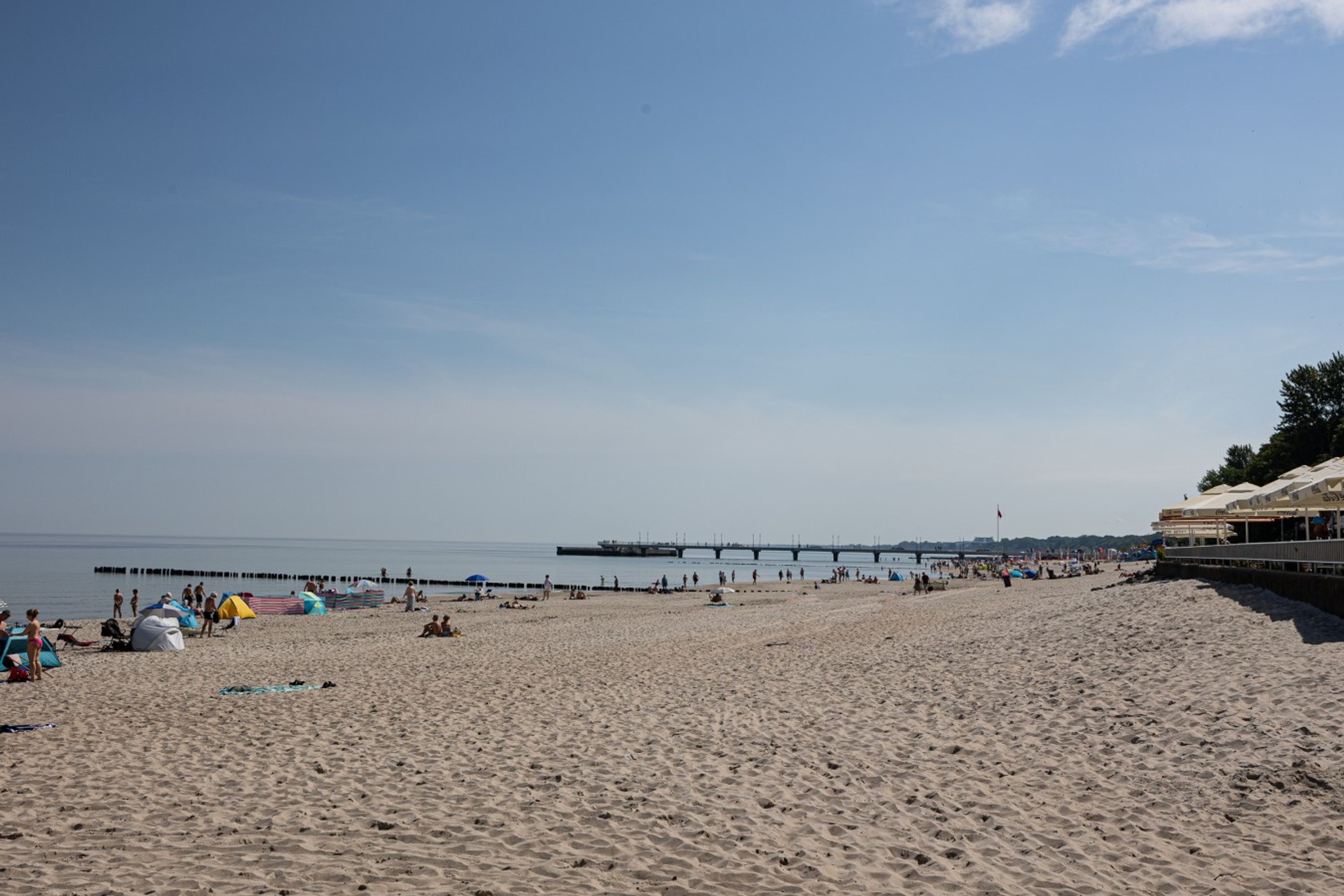Una playa con un muelle que se adentra en el agua, arena y un edificio con un área cubierta a la derecha.