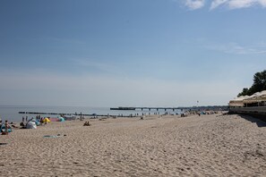 Ein Strand mit einem Steg, der sich ins Wasser erstreckt, feinem Sand und einem Gebäude mit überdachtem Bereich rechts.
