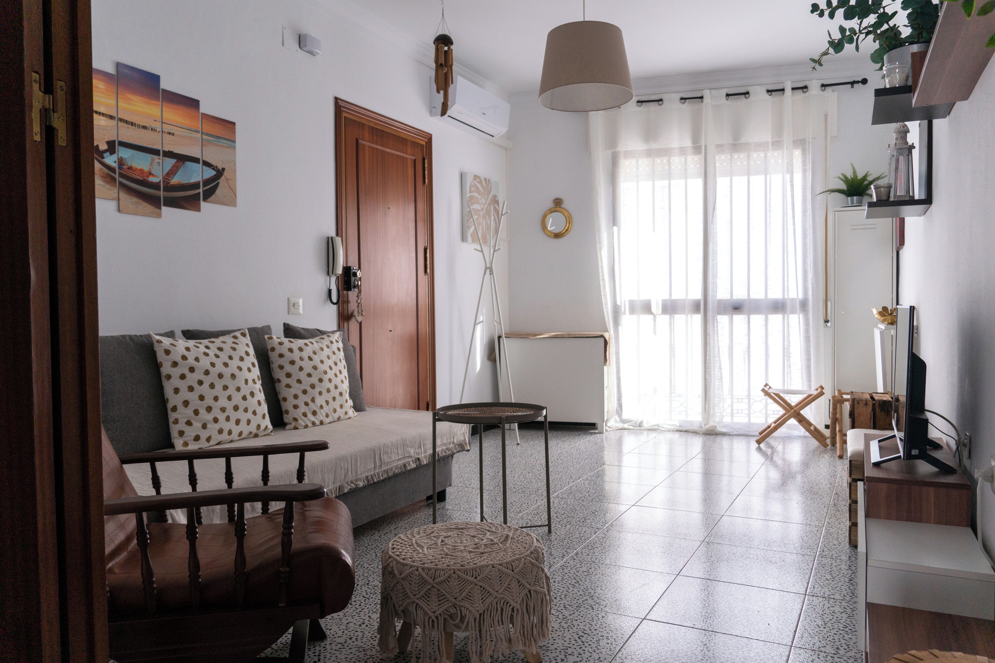 Uma sala de estar com sofá, mesa de centro, mesa redonda pequena, cadeira de madeira, banquinho estampado, televisão e uma janela grande com cortinas leves.