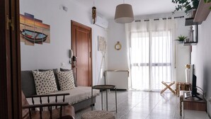 Ein Wohnzimmer mit einer Couch, einem Couchtisch, einem kleinen runden Tisch, einem Holzstuhl, einem gemusterten Hocker, einem Fernseher und einem großen Fenster mit durchsichtigen Vorhängen.