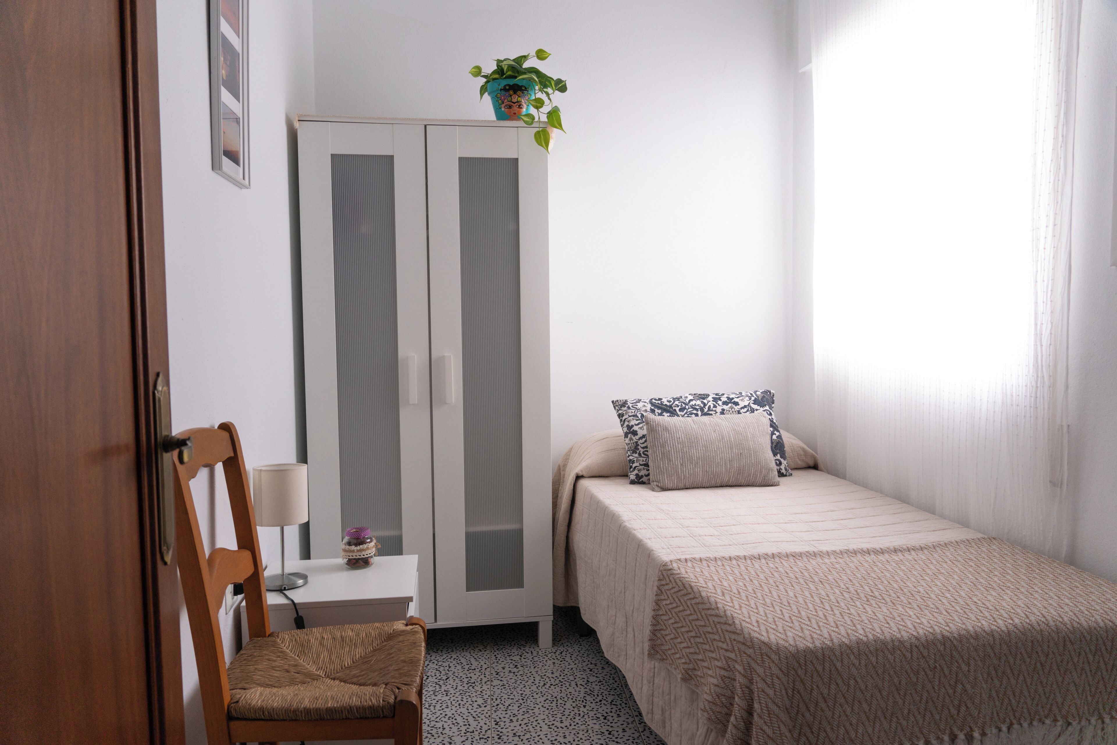 Un dormitorio con cama, una silla, armario, escritorio con lámpara y planta en maceta.